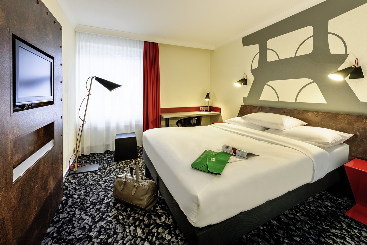 Modernes Hotelzimmer mit Doppelbett, roter Wand, grauen Teppichboden und heller Beleuchtung.