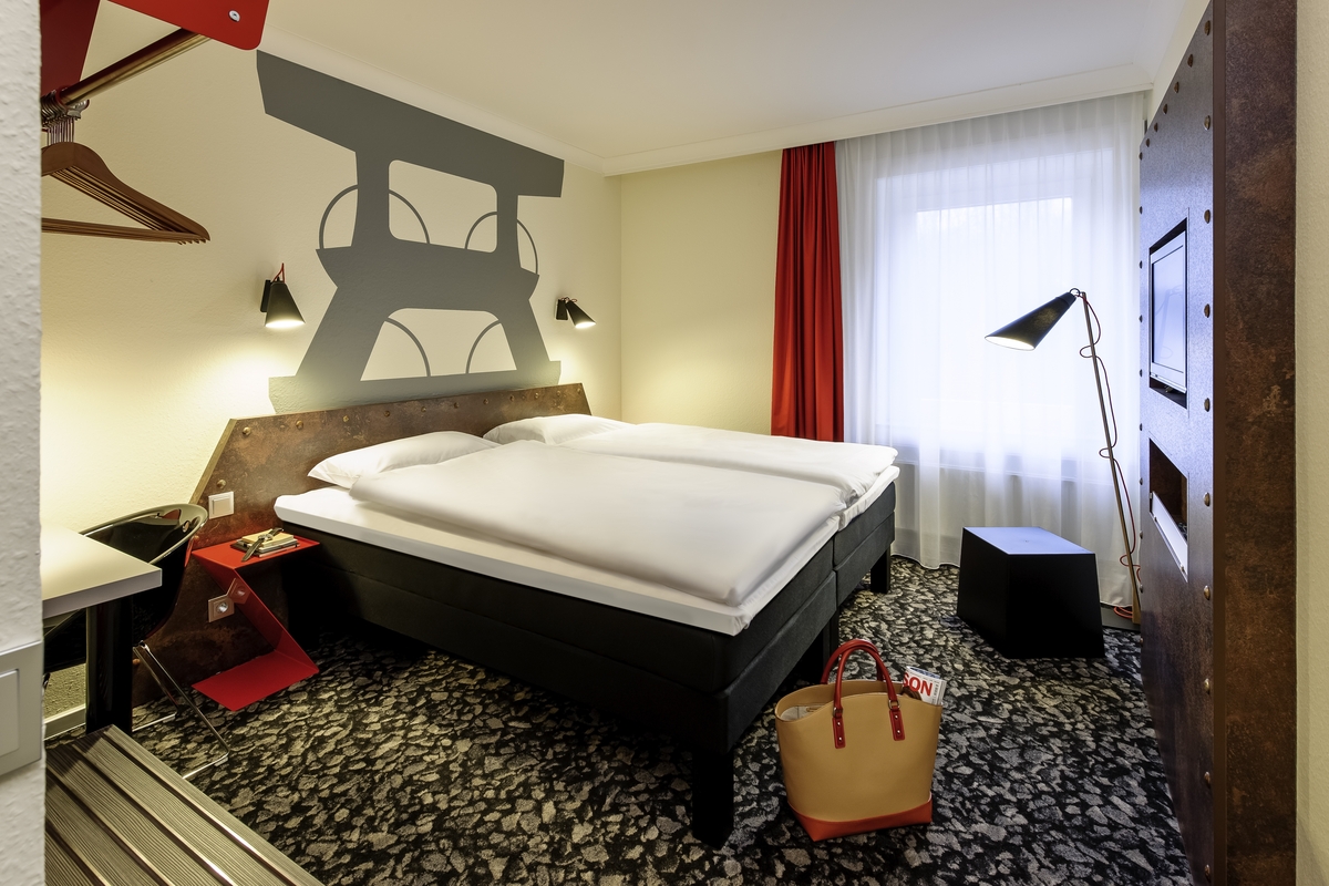 Moderner Hotelraum mit Doppelbett, roten Akzenten und Wandkunst. Helle Beleuchtung. Keine Personen.