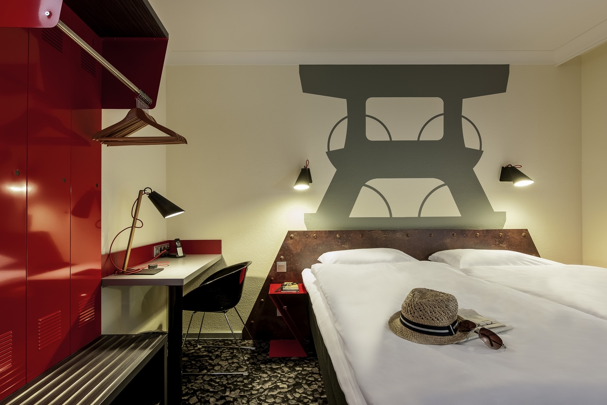 Ein modernes Hotelzimmer mit Doppelbett, roter Schrank, Strohhut auf Bett und Wandkunst.