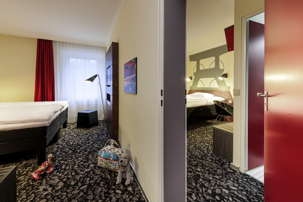 Ein Hotelzimmer mit zwei Betten, bunter Tasche, Kuscheltier und rotem Badezimmertürrahmen.