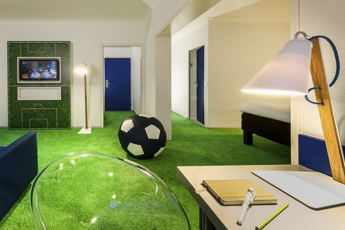 Raum mit grünem Teppich, Fußballthemen-Dekor, großem Fußball, blauem Sofa und beleuchtetem Tisch.