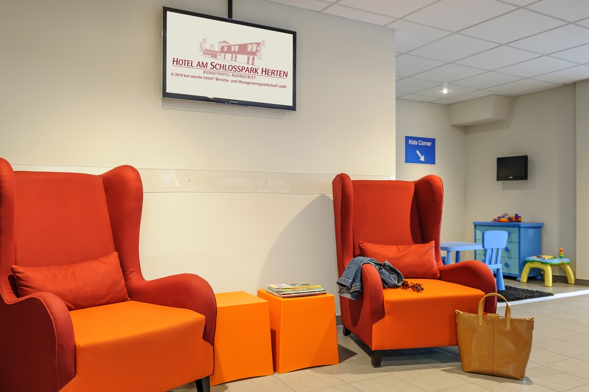 Zwei rote Sessel in einer Hotellobby mit Kinderspielecke und einer orangefarbenen Tasche.