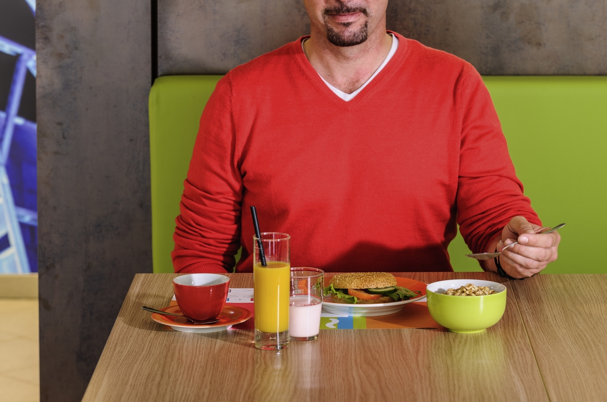 Mann in rotem Pullover sitzt lächelnd am Tisch mit Saft, Kaffee, Müsli und Burger.