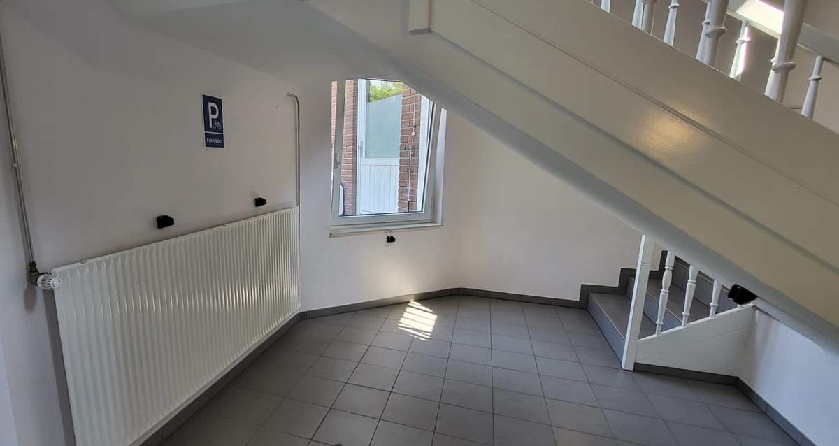 Treppenhaus mit weißen Wänden, grauen Fliesen, Heizkörper, Fenster und Parkplatzschild.