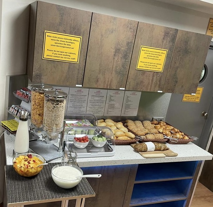 Frühstücksbuffet mit Cerealien, Brötchen, Obst und Milch; Holzmöbel; Infotafeln an Wänden.