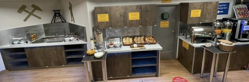 Ein Frühstücksbuffet mit Brötchen, Müsli, Saftspendern und Kaffeemaschinen in einem modernen Raum.