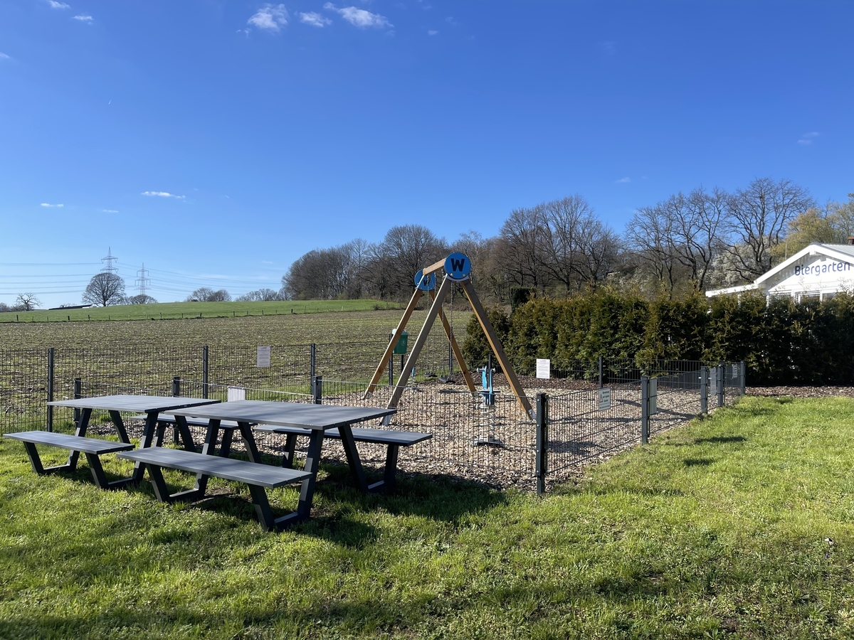 Spielplatz mit Schaukel, Holztische im Vordergrund, blauer Himmel, Wiese und Bäume im Hintergrund.
