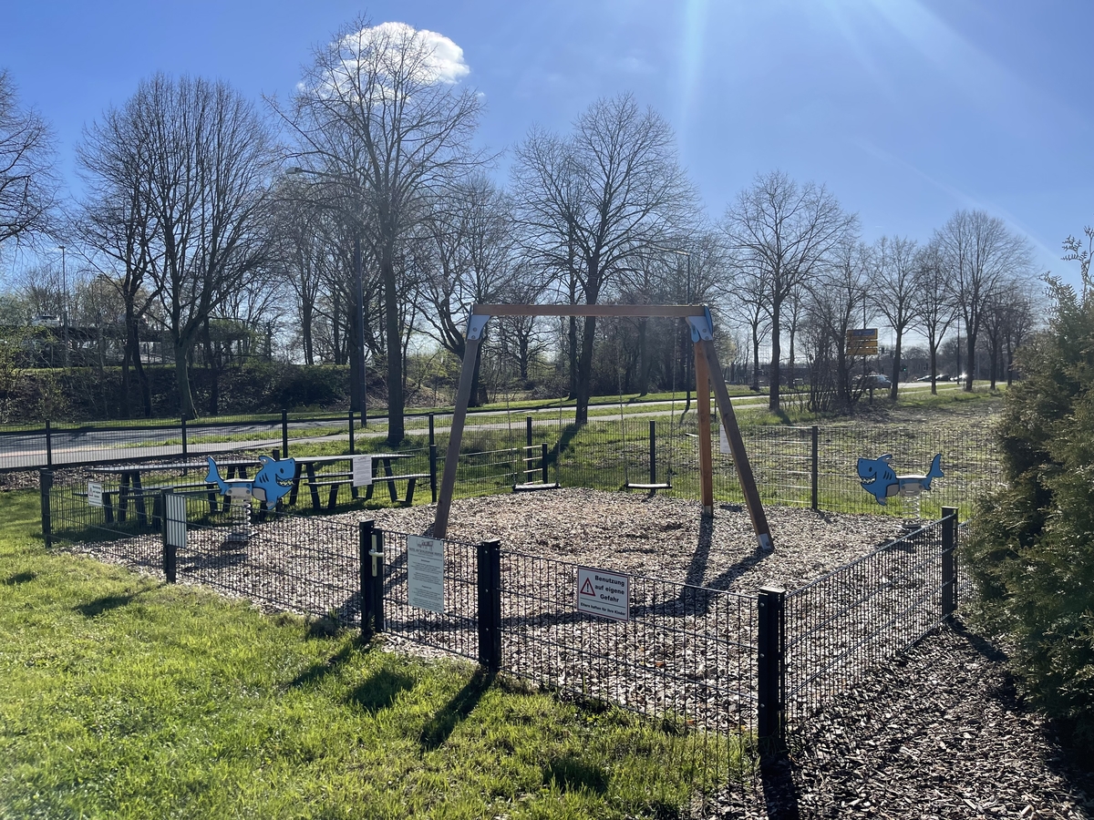 Spielplatz mit Wippe, Schaukeltier und Bänken auf Holzboden. Bäume und blauer Himmel im Hintergrund.