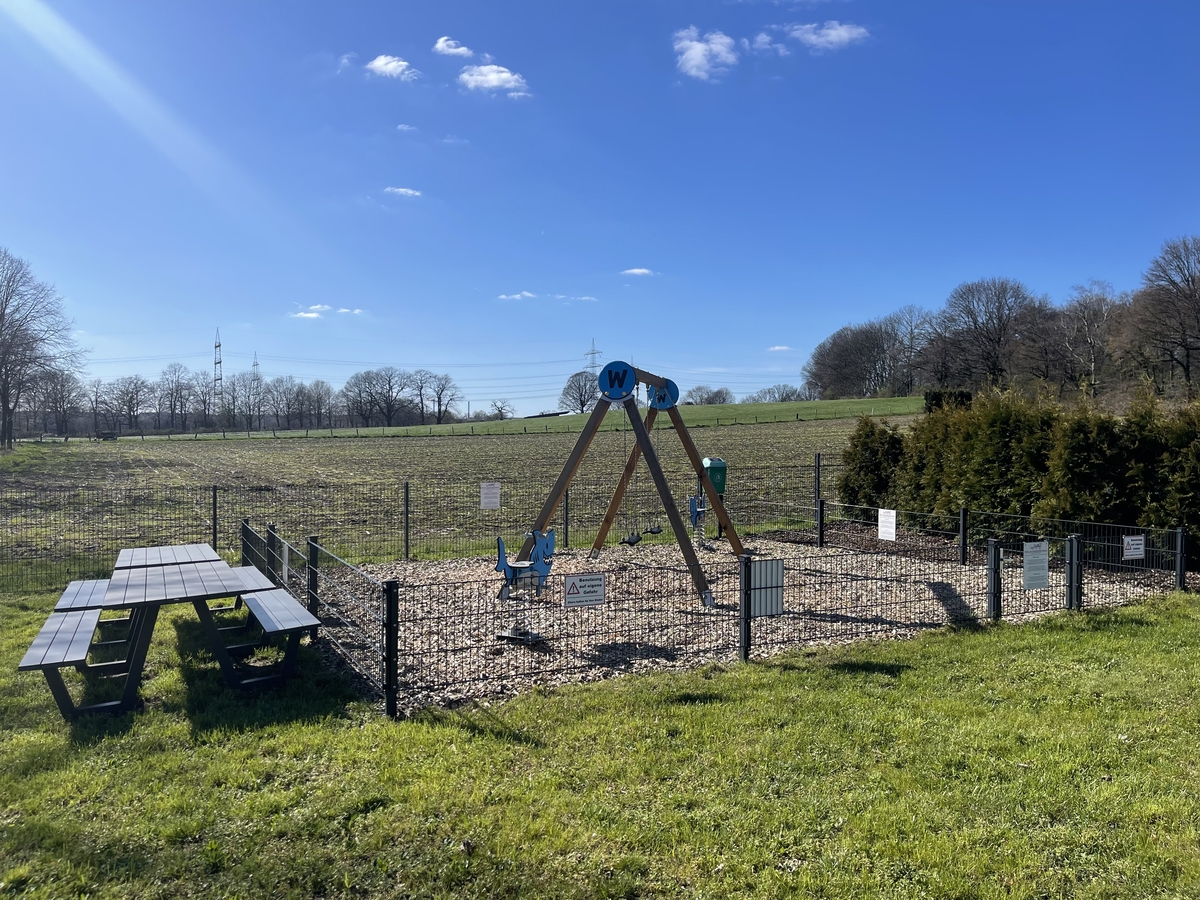 Ein Spielplatz mit Schaukeln, umgeben von Wiesen unter blauem Himmel an einem sonnigen Tag.