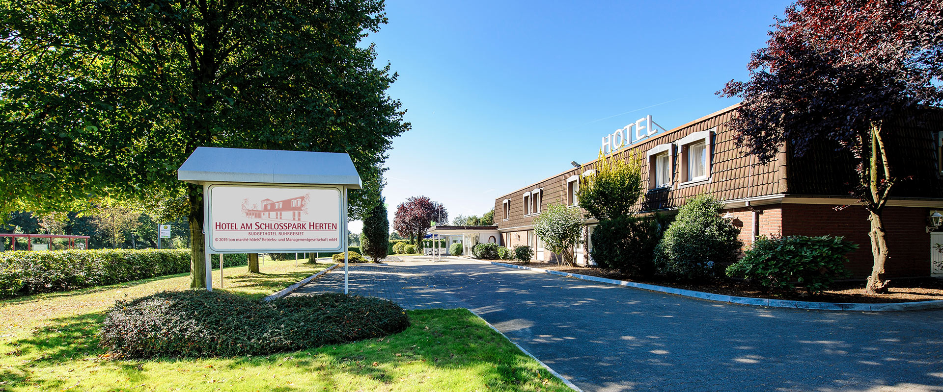 Hotelfassade bei Tageslicht, umgeben von Bäumen, mit großem Werbeschild im Vordergrund.
