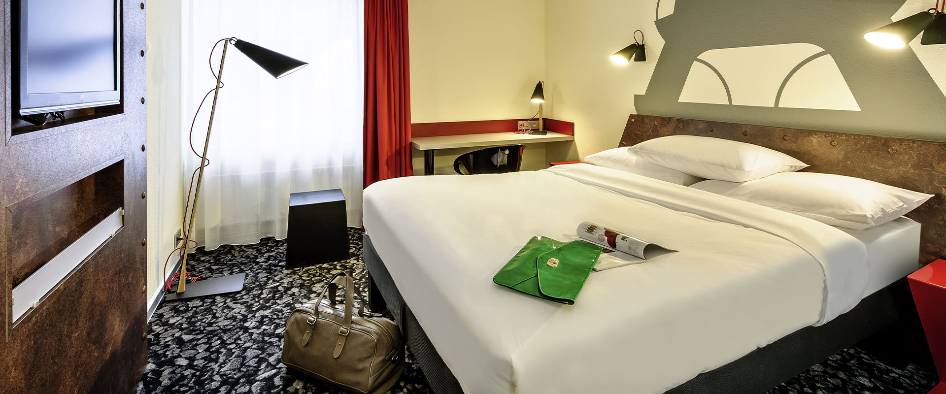 Modernes Hotelzimmer mit einem großen Bett, Ledertasche, Tischlampe und abstrakter Wandkunst.